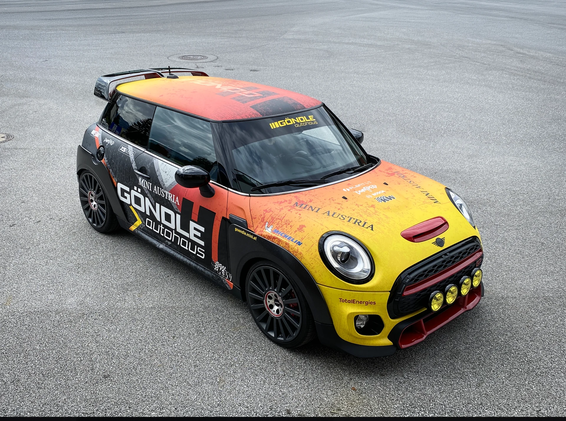 Motorsport Verlauf Folierung MINI Autohaus Göndle – Fahrzeugdesign mit Farbverlauf in Gelb und Rot, mit Stickern Dumped, KW, XS MAG, Michelin